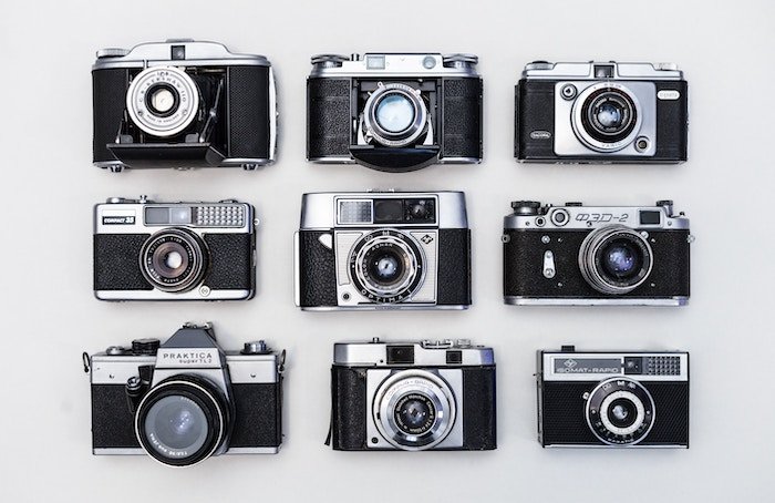 Nine silver and black cameras arranged in neat lines on a table and shot from above